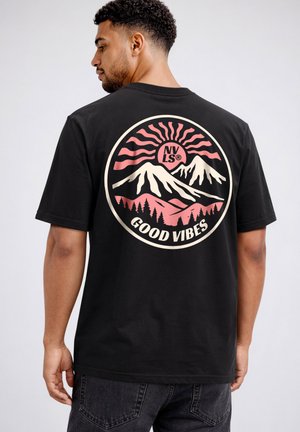 Man wearing black t-shirt with circular pink and cream mountain, sun, trees design, and text "NVLS® GOOD VIBES" on the back.