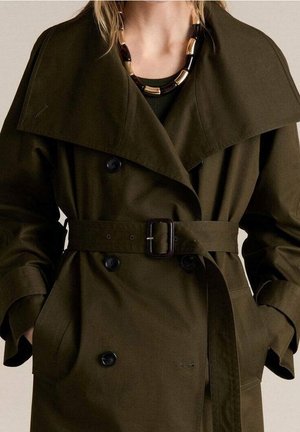 Femme portant un trench-coat ceinturé vert olive foncé avec de larges revers et un collier épais noir et or, mains dans les poches.