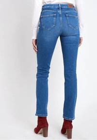 Pantalones vaqueros de mezclilla azul oscuro con un corte ajustado, que cuentan con bolsillos traseros, una cintura de altura media y un sutil efecto de desgaste. Combinados con botines de tacón rojos.