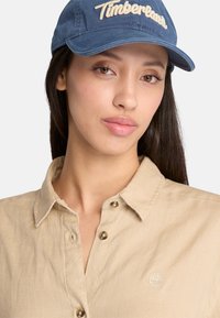Camicia beige con bottoni e colletto, con logo Timberland. Cappellino blu scuro con ricamo della scritta "Timberland", visiera strutturata. Tessuto liscio.