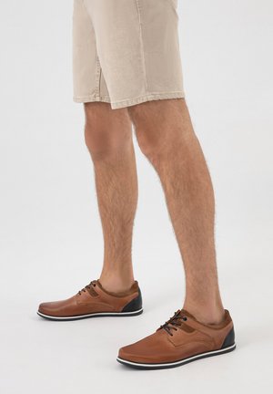 Uomo con pantaloncini beige e scarpe casual di pelle marrone con lacci, in piedi su un pavimento grigio chiaro.