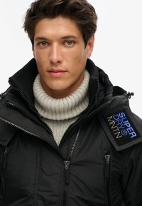 Zwarte, ritssluitingjack met geribbelde kraag en ritszakken, voorzien van een textuurstof en geborduurd "SUPERDRY MNTN"-label op de schouder.