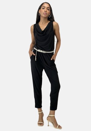 Zwarte jumpsuit met een gedrapeerde halslijn, elastische taille, zakken en een gevlochten witte riem. Gdrag met strappy tan hakschoenen.