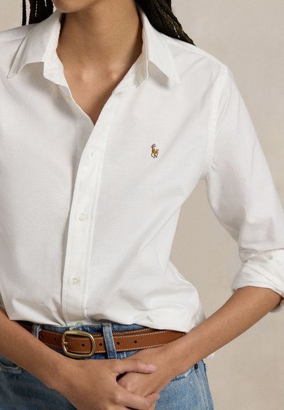 Person som har på seg en hvit skjorte med knapper og en liten brodert hestelogo, stukket inn i blå jeans med et brunt lærbelte.