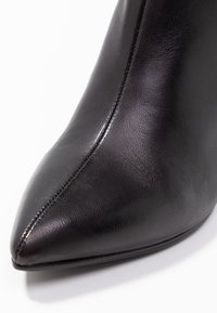 Stivaletto in pelle nera con punta appuntita, texture liscia, cuciture minimali e silhouette elegante, posato su uno sfondo bianco.