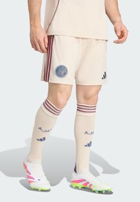 Mężczyzna ubrany w kremowe spodenki piłkarskie Ajax, dopasowane skarpetki w bordo paski oraz białe korki z neonowo-zielonymi i różowymi akcentami.