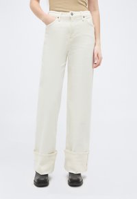 Personne portant un pantalon blanc à jambes larges et taille haute avec de grands revers, par-dessus des chaussures noires, debout avec une main dans la poche sur un fond blanc.