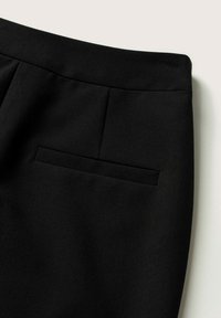 Pantalon noir sur mesure avec une ceinture plate et une unique poche passepoilée horizontale à l’arrière sur un fond neutre.
