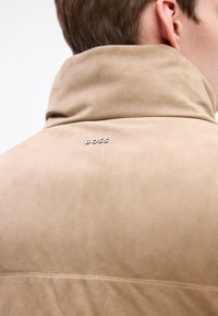 Beige ruskindsjakke med høj krave, prydet med et diskret "BOSS" logo i sølv på ryggen. Glat tekstur og minimalistisk design.