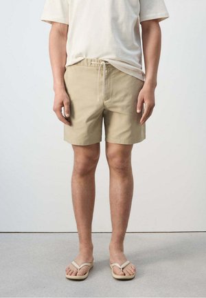 Persona con pantalones cortos beige con cordón, camiseta blanca y chanclas beige, de pie sobre suelo gris frente a una pared lisa.