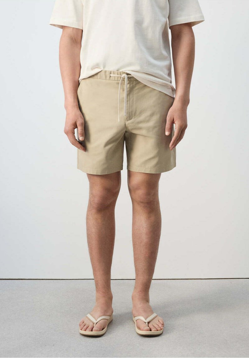 Personne portant un short beige avec cordon, un t-shirt blanc et des tongs beiges, debout sur un sol gris devant un mur uni.