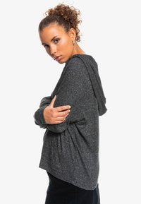 Mørkegrå hættetrøje lavet af blødt, struktureret stof. Har en hætte, lange ærmer og en afslappet pasform med sideslidser for komfort.