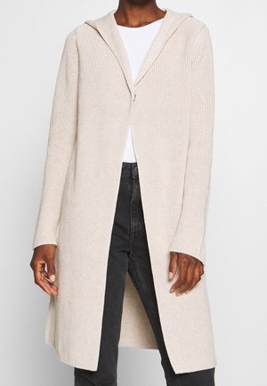 Personne portant un long cardigan tricoté beige clair avec une capuche, par-dessus une chemise blanche et un pantalon foncé, debout devant un fond uni.