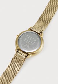 Montre avec bracelet en maille dorée, présentant un boîtier rond, une finition métallique et un dos gravé avec le logo et les détails de la marque. Résistante à l'eau jusqu'à 3 bars.