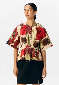 Red and cream patterned short-sleeve blouse with a gathered neckline and adjustable drawstring waist, paired with a black skirt.