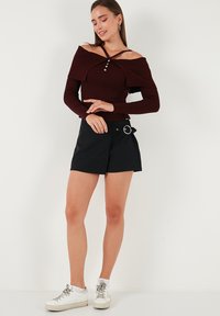 Raudonas ribotas off-shoulder megztinis su dekoratyvinėmis sagomis, derinamas su juodais apvyniojimo šortais, turinčiais didelį akcento diržo sagtį ir baltais sportbačiais.