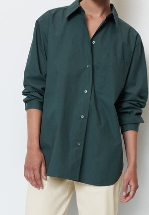 Mand iført en mørkegrøn skjorte med knapper og opkørte ærmer samt beige bukser, stående mod en ensfarvet hvid baggrund.