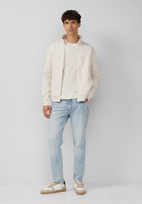 Licht crème zip-up jasje over een wit shirt, gecombineerd met lichtblauwe jeans en witte sneakers met beige en marineblauwe accenten. Soepele stofstructuur.