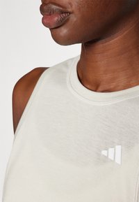 Jasnobeżowy, bezrękawnik wykonany z gładkiego materiału, z białym logo przedstawiającym trzy trójkątne kształty po lewej stronie.