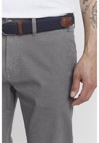 Pantalones de tela gris tejido con una superficie texturizada, acompañados de un cinturón trenzado marino con un acento de cuero marrón. Primer plano de la zona de la cintura.