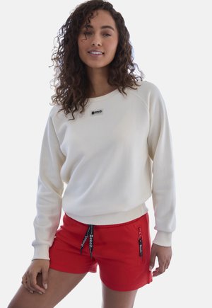 Woman with curly hair wearing a white long-sleeve sweatshirt and red shorts with black drawstrings and zipper pocket details.
