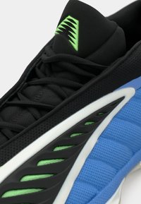 Zapatilla deportiva negra con un upper texturizado, una media suela azul y blanca, detalles de acento verdes y un diseño de lengüeta distintivo con patrón.