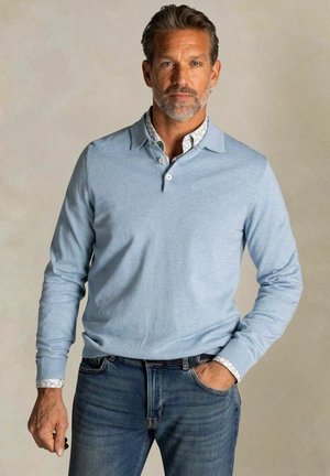 Man met grijs haar en baard, draagt een lichtblauw poloshirt met lange mouwen over een gebloemd overhemd, blauwe spijkerbroek, met één hand in de zak, neutrale achtergrond.