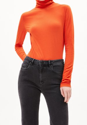 Kvinde iført en tætsiddende, lys orange langærmet rullekrave, stoppet ned i sorte højttaljede jeans mod en hvid baggrund.