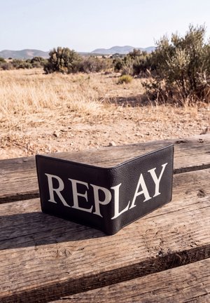 Una cartera negra de cuero con la marca REPLAY está sobre un banco de madera desgastado en un paisaje seco cubierto de arbustos.
