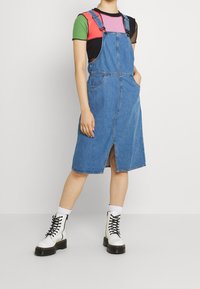 Jeansbyxdress med justerbara axelremmar, framfickor och en mittslits; matchad med en randig kortärmad topp och vita ankelskor.