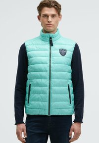 Giovane uomo che indossa un gilet trapuntato blu chiaro senza maniche con tasche con zip su una camicia nera a maniche lunghe, in piedi davanti a uno sfondo bianco puro.