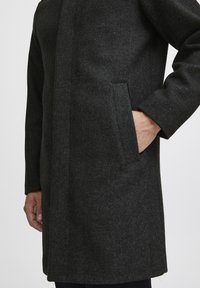 Mørk grå ullfrakk med teksturert overflate. Har en rett skjæring, sidelommer og et design uten krage. Nærbilde av stoffet og passformen.