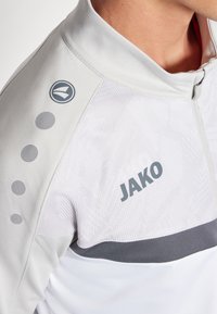 Jachetă sport de culoare gri deschis, cu guler cu fermoar, având un model texturat, accente negre și branding "JAKO" pe piept și umăr.