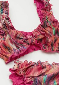 Barevný bikiny top s volánovými okraji, vyrobený ze lesklé látky se spirálovým vzorem v růžové, oranžové a modré barvě. Obsahuje detail mašle vpředu.