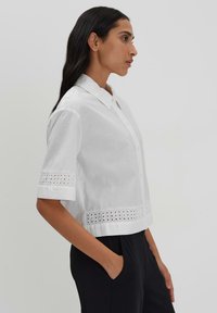 White cropped shirt with short sleeves, featuring a button-down front and decorative eyelet trim along the sleeves and hem.