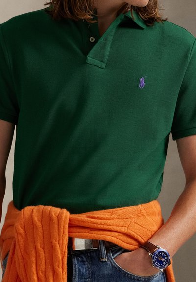 Osoba u zelenoj polo majici s ljubičastim logom, narančastom džemperu vezanom oko struka, plavim trapericama i plavim ručnim satom s smeđom kožnom narukvicom.