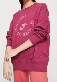 Vinröd sweatshirt med en stor rosa Nike-logotyp och grafisk text, med ribbade mudd och nederkant, tillverkad av mjukt, texturerat material.