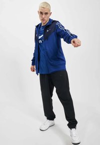 Zila kapucēta džemperis ar melniem akcentiem, uzvilkts virs zila T-kreklu. Melnas sporta bikses ar logotipa detaļām, kombinētas ar baltām sporta apaviem.