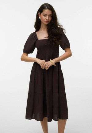 Hosszú sötét hajú nő sötétbarna midi ruhát visel, puffos ujjal, négyzet alakú nyakkivágással és réteges szoknyával, sima háttér előtt állva.