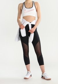 White sports bra with black trim and red logo, paired with black leggings featuring mesh panels and white zip-up top tied around the waist.