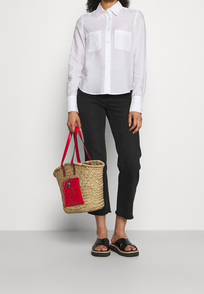 Personne portant une chemise blanche boutonnée et un pantalon noir raccourci, tenant un sac en paille tressée avec des poignées rouges et une poche, portant des sandales noires.