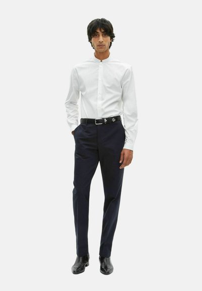Man staat in een witte mandarin kraag shirt, zwarte riem, marineblauwe broek en zwarte nette schoenen op een witte achtergrond.