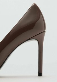 Pantof din piele lacuită maro, cu toc înalt, vârf ascuțit și siluetă elegantă. Are un toc stiletto subțire și un finisaj neted și strălucitor.