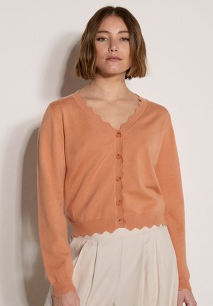 Giovane donna che indossa un cardigan pesca con bordi a smerlo e bottoni, e pantaloni beige chiaro a vita alta, su uno sfondo semplice.