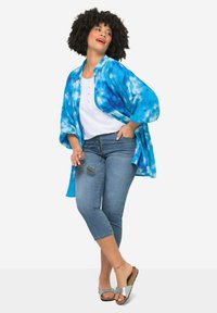Cardigan azul claro con diseño de tie-dye, camisa blanca con botones y jeans de mezclilla cortos. Modelo lleva sandalias plateadas y sostiene unas gafas de sol.