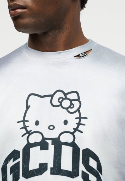 Mies, joka pukeutunut valkoiseen paitaan, jossa musta Hello Kitty -kasvokuvio ja GCDS-logo, pienen leikkauksen kanssa kaula-aukon lähellä, jossa on GCDS-etiketti.