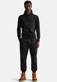 Czarna bluza z kapturem z wytłoczonym logo, pasujące dresy oraz pomarańczowe buty z czarnymi akcentami i detalami sznurowania, wszystko wykonane z miękkiej, komfortowej tkaniny.