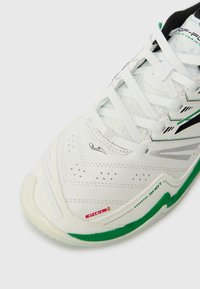 Białe buty sportowe z zielonymi i czarnymi detalami, białymi sznurówkami i teksturowaną podeszwą, z widocznymi logotypami marki oraz nazwą modelu.