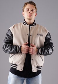 Beige Wolle-Mix Varsity-Jacke mit schwarzen Lederärmeln, schwarzen gestreiften Rippbündchen und einem Logo auf der Brust. Druckknopfverschluss.