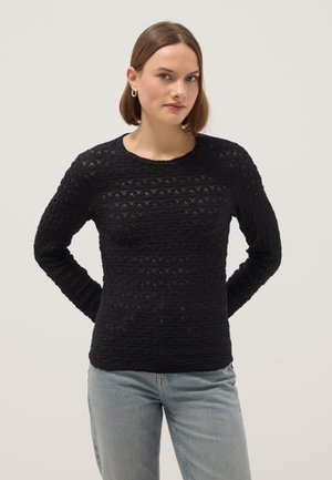 Donna con capelli corti castani che indossa orecchini a cerchio dorati, una maglia nera a maniche lunghe con texture e jeans blu chiaro su uno sfondo semplice.